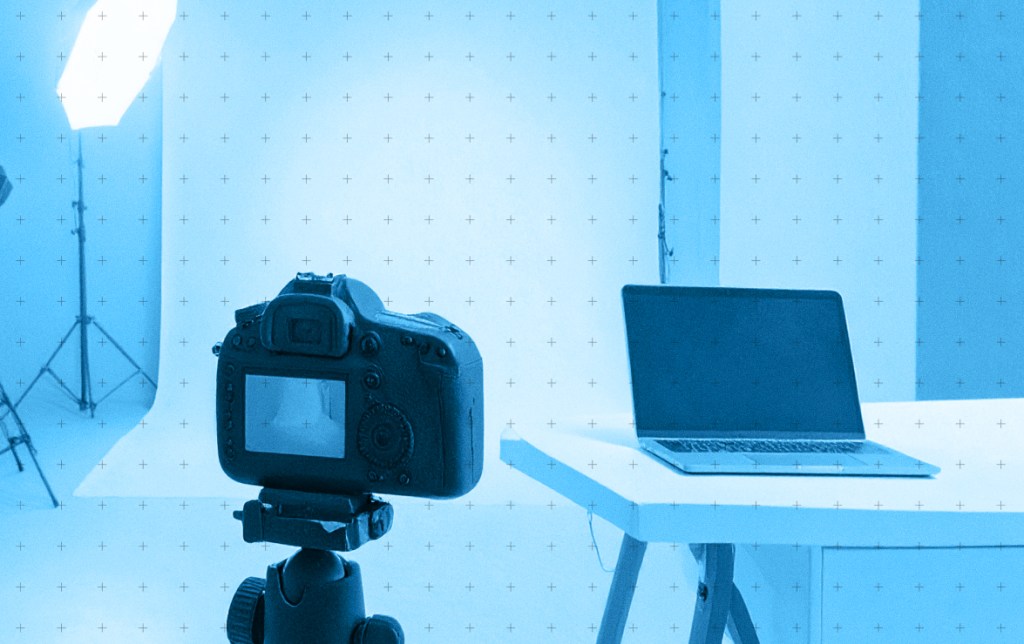 A professional camera set up on a tripod facing a laptop on a table, with a bright, softly lit backdrop suitable for photography.